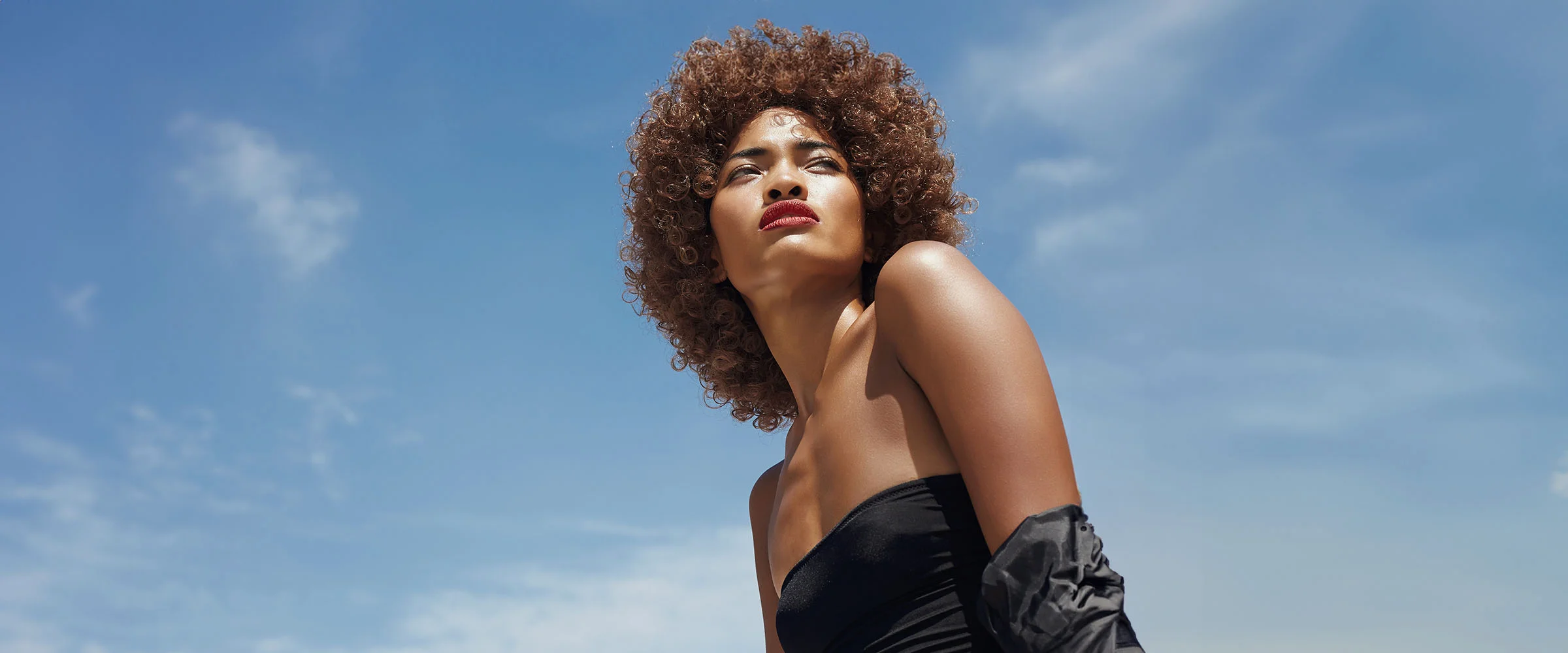 A woman with curly hair and red lipstick, dressed in a black strapless top, gazing upward against a clear blue sky - Laser Resurfacing Procedure in New York, NY