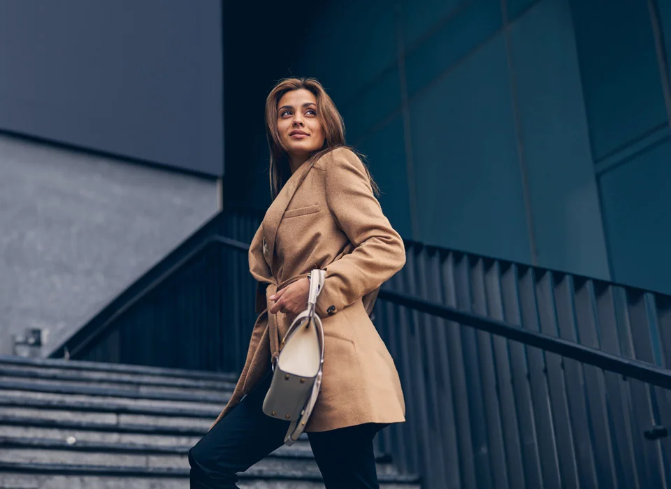 A stylish woman wearing a camel coat and carrying a white handbag, ascending a flight of stairs outside a building - Out of Town patient resources in New York City