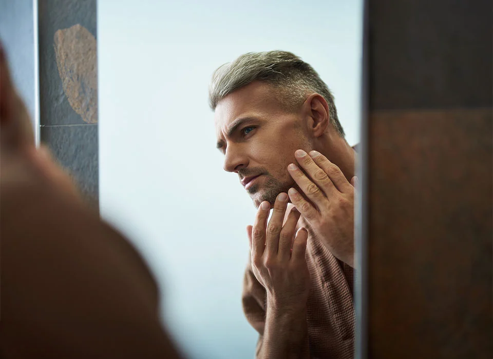 A white haired man touching his face, while looking at his reflection in the mirror - Face Lift Procedure in New York, NY